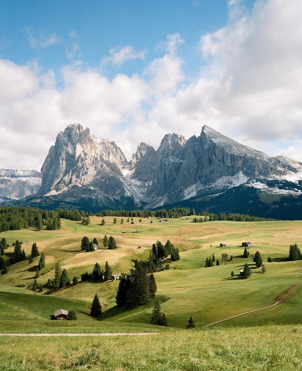 A vast green valley with scattered trees and small wooden huts stretches towards a range of snow-capped mountains under a partly cloudy sky.