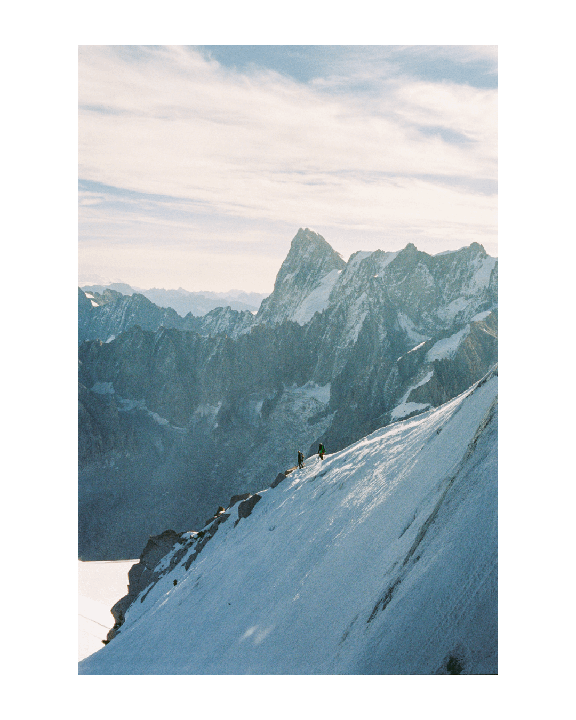 Two people are hiking up a snow-covered mountain slope with a mountain range in the background.