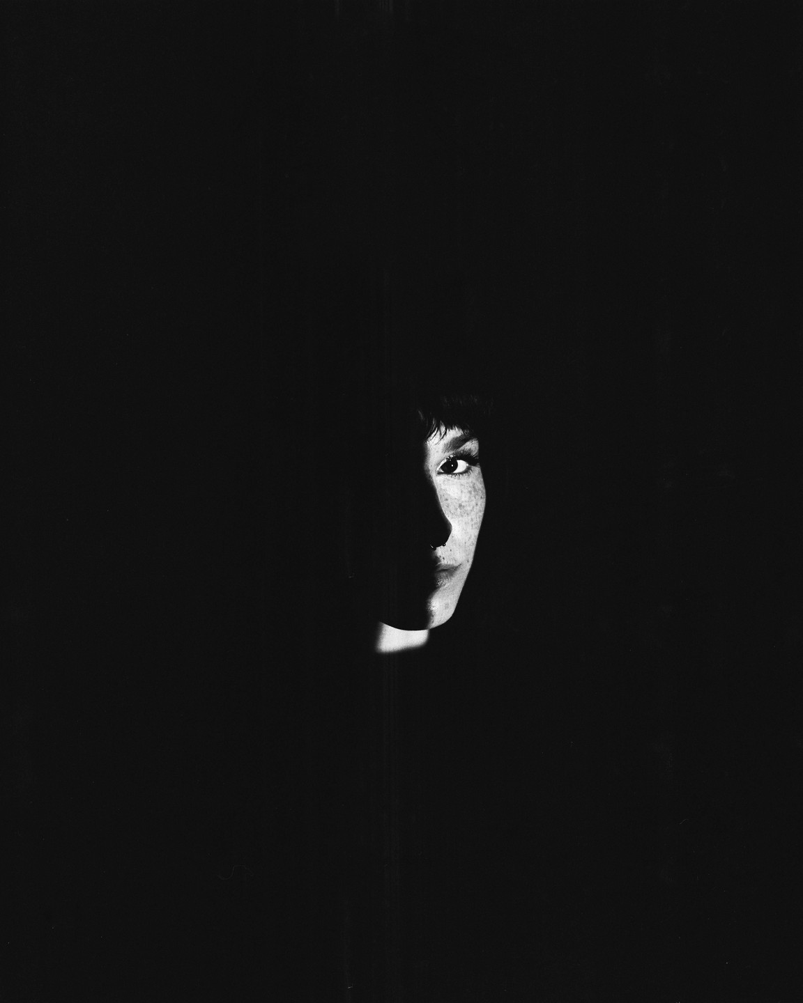 A woman's face is partially illuminated by a sliver of light in a dark setting.