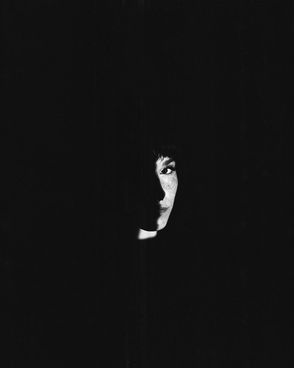 A woman's face is partially illuminated by a sliver of light in a dark setting.