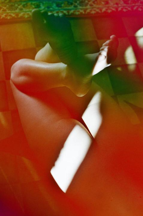 A person's legs and feet are visible on a tiled floor with colorful light leaks and strong shadows.