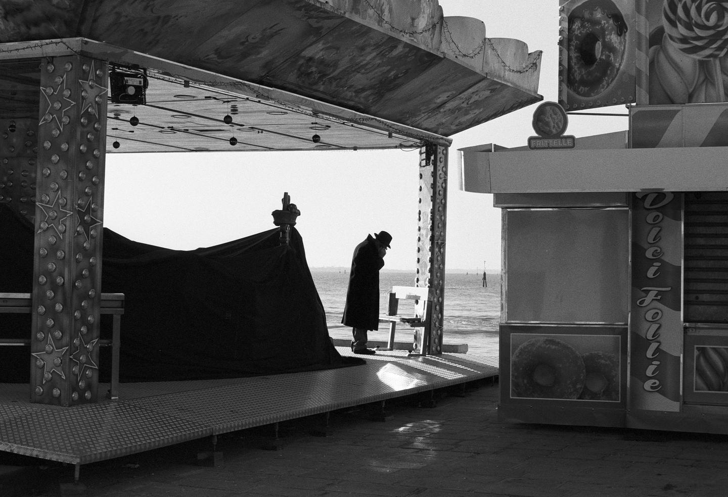 A man in a hat stands by a covered stall next to the sea.