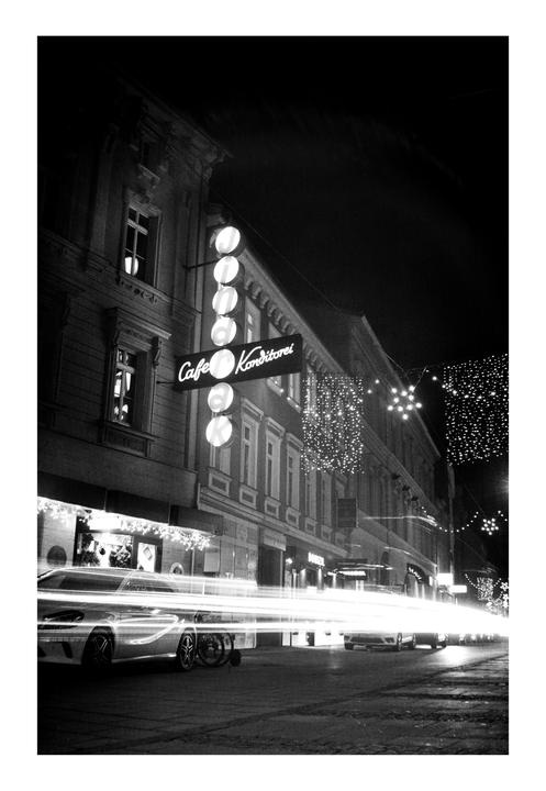 A street at night is illuminated by a neon sign for a cafe and the light trails of passing cars.