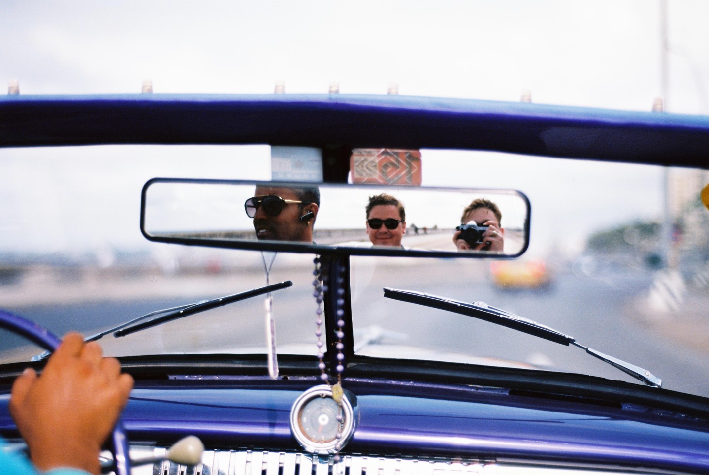 Three men are visible in the rearview mirror of a purple car, with one man holding a camera.