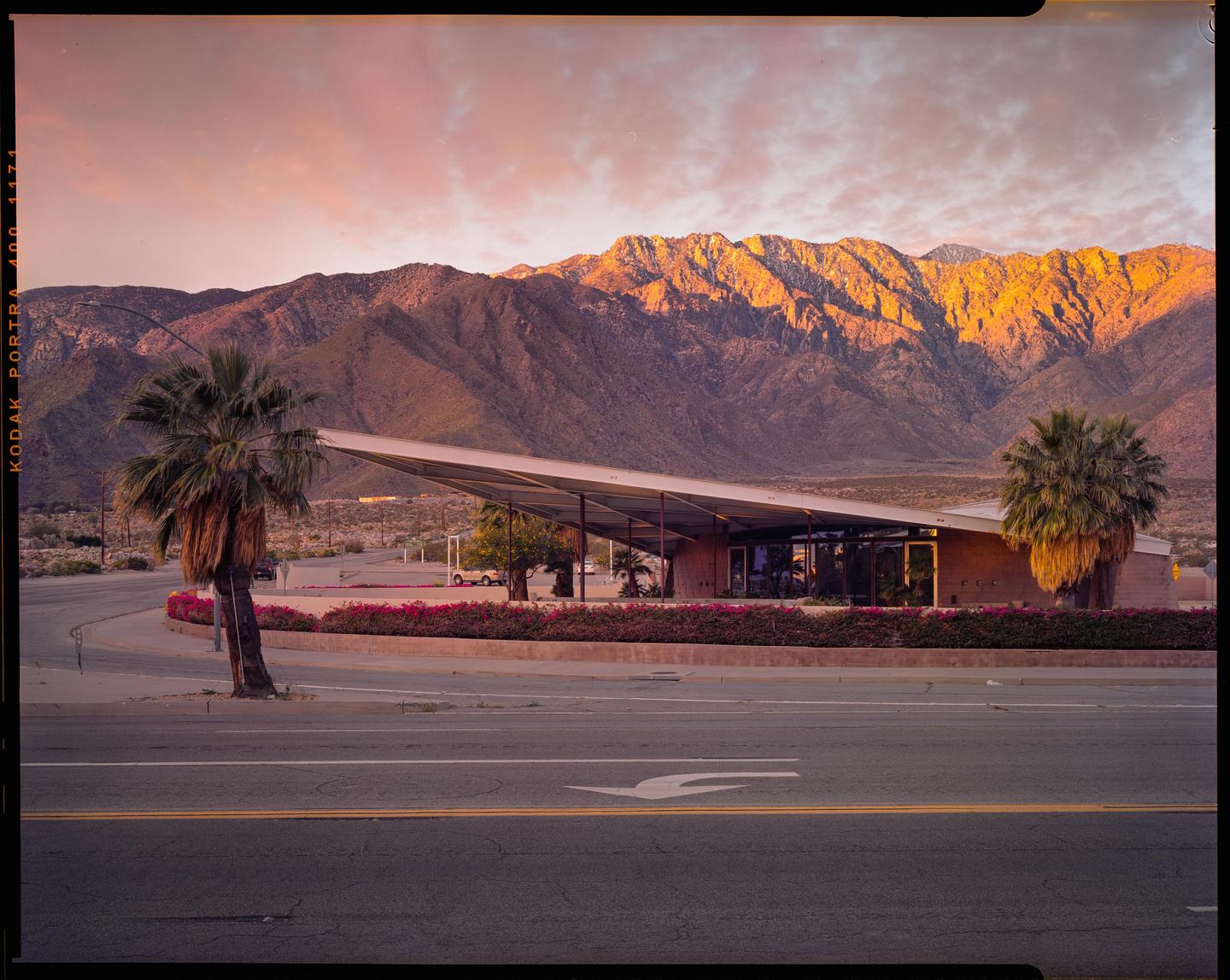 A modern building with a large angled roof stands in front of a mountain range under a colorful sky.