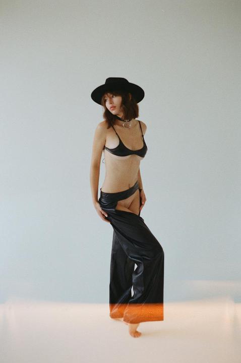 A woman wearing a black hat, bra, and chaps stands against a light background with an orange light leak at the bottom.