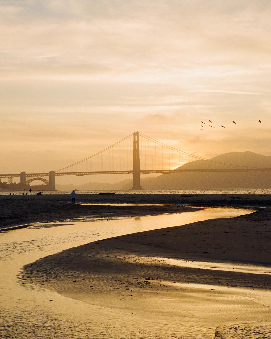 A golden hour view of the Golden Gate Bridge from a sandy beach with a winding stream.