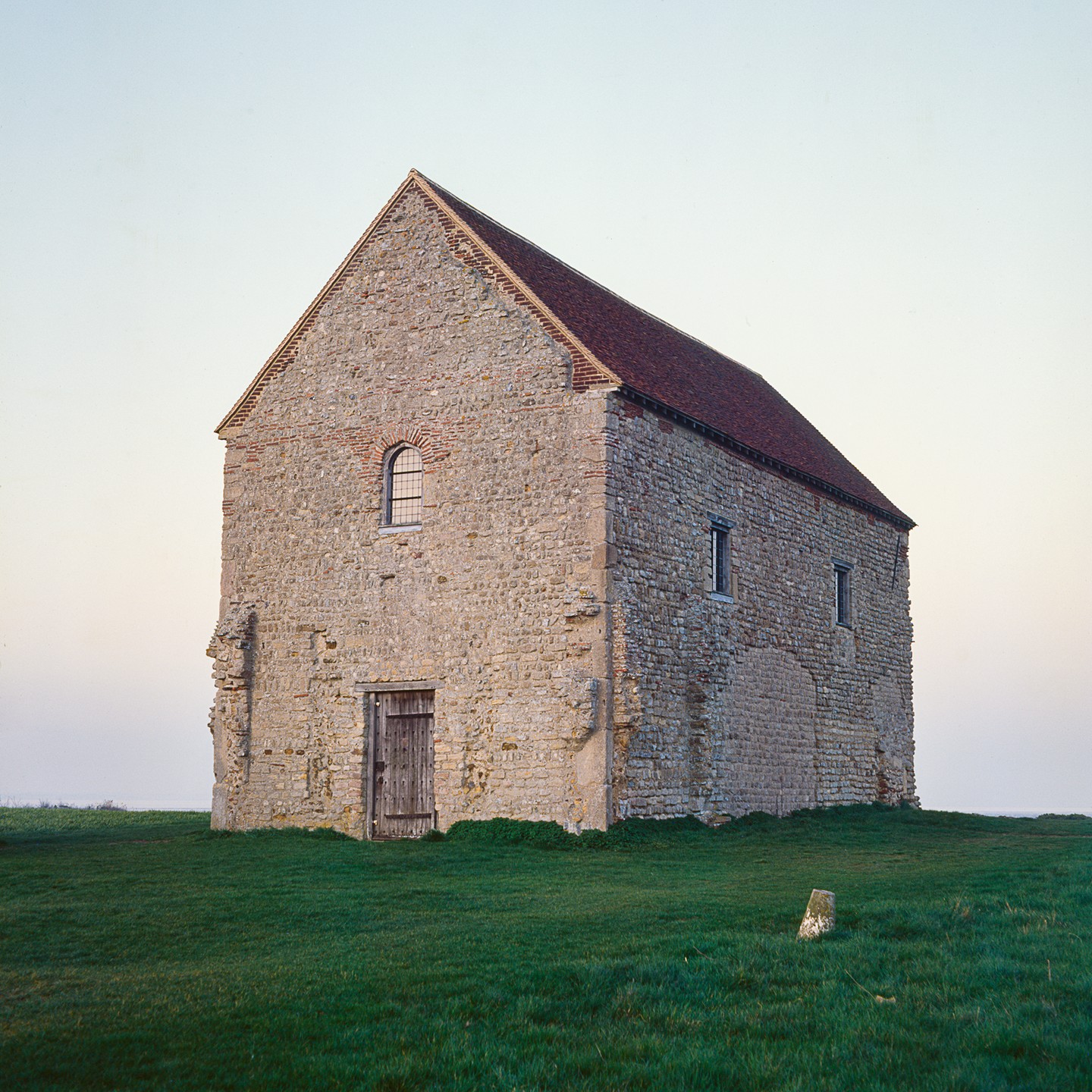 A stone church with a red tile roof stands on a grassy hill under a pale sky.