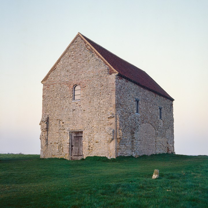 A stone church with a red tile roof stands on a grassy hill under a pale sky.
