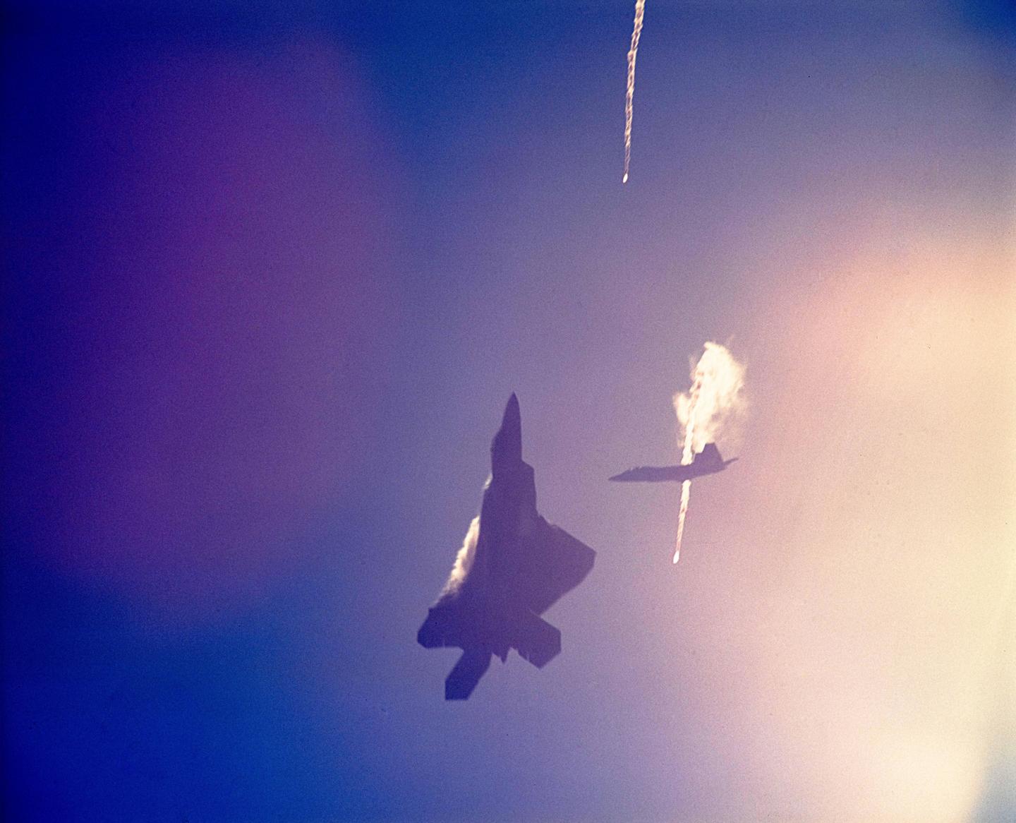 Two fighter jets are visible in the sky, one with a trail of smoke and the other with a bright light on its underside.