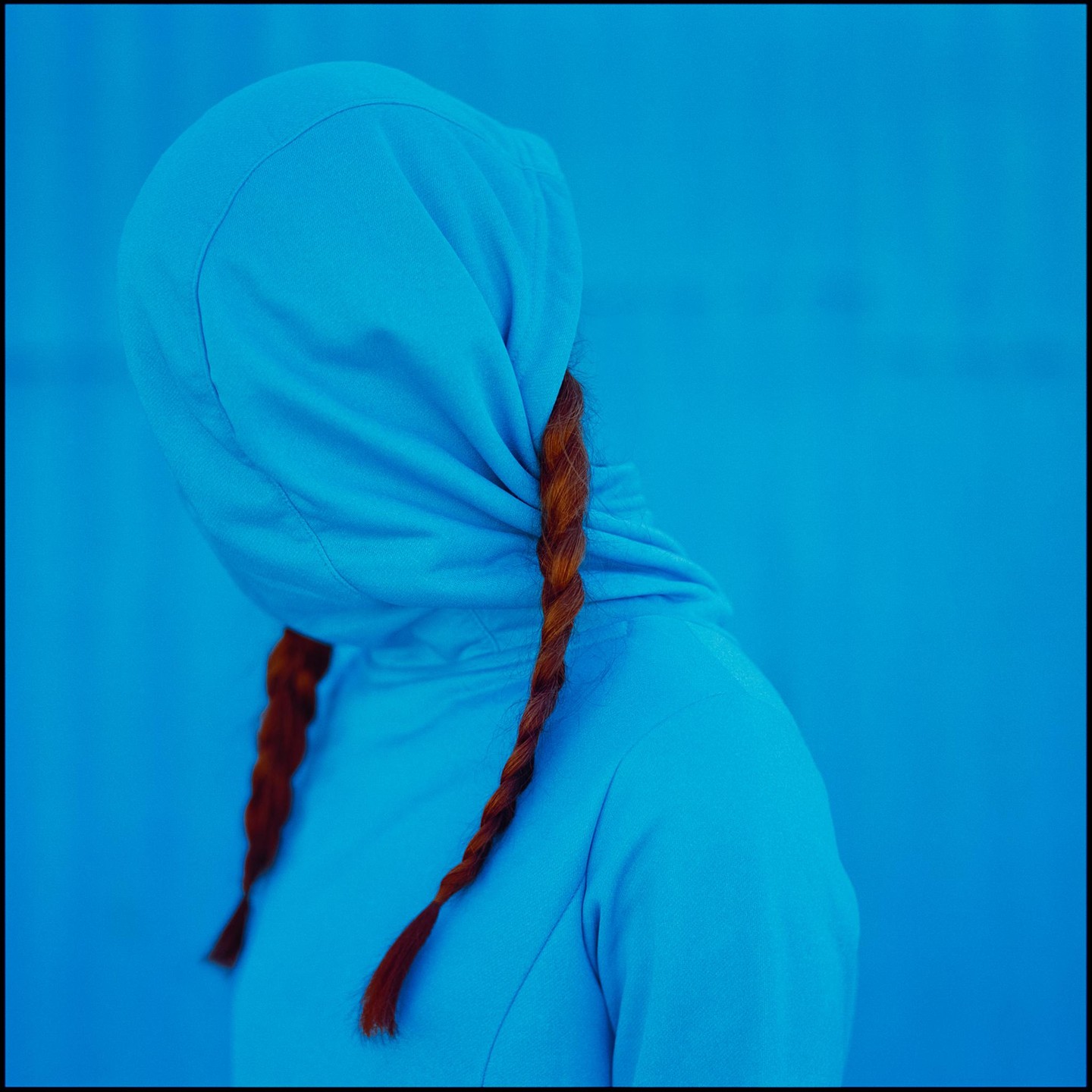 A person with braided red hair wears a blue hooded sweatshirt against a blue background.