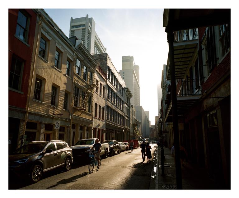 A person rides a bicycle down a sunlit street lined with buildings and parked cars in New Orleans.