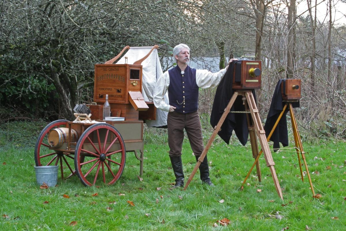 A man in period clothing stands next to a large format camera on a tripod and a mobile darkroom cart in a grassy area.