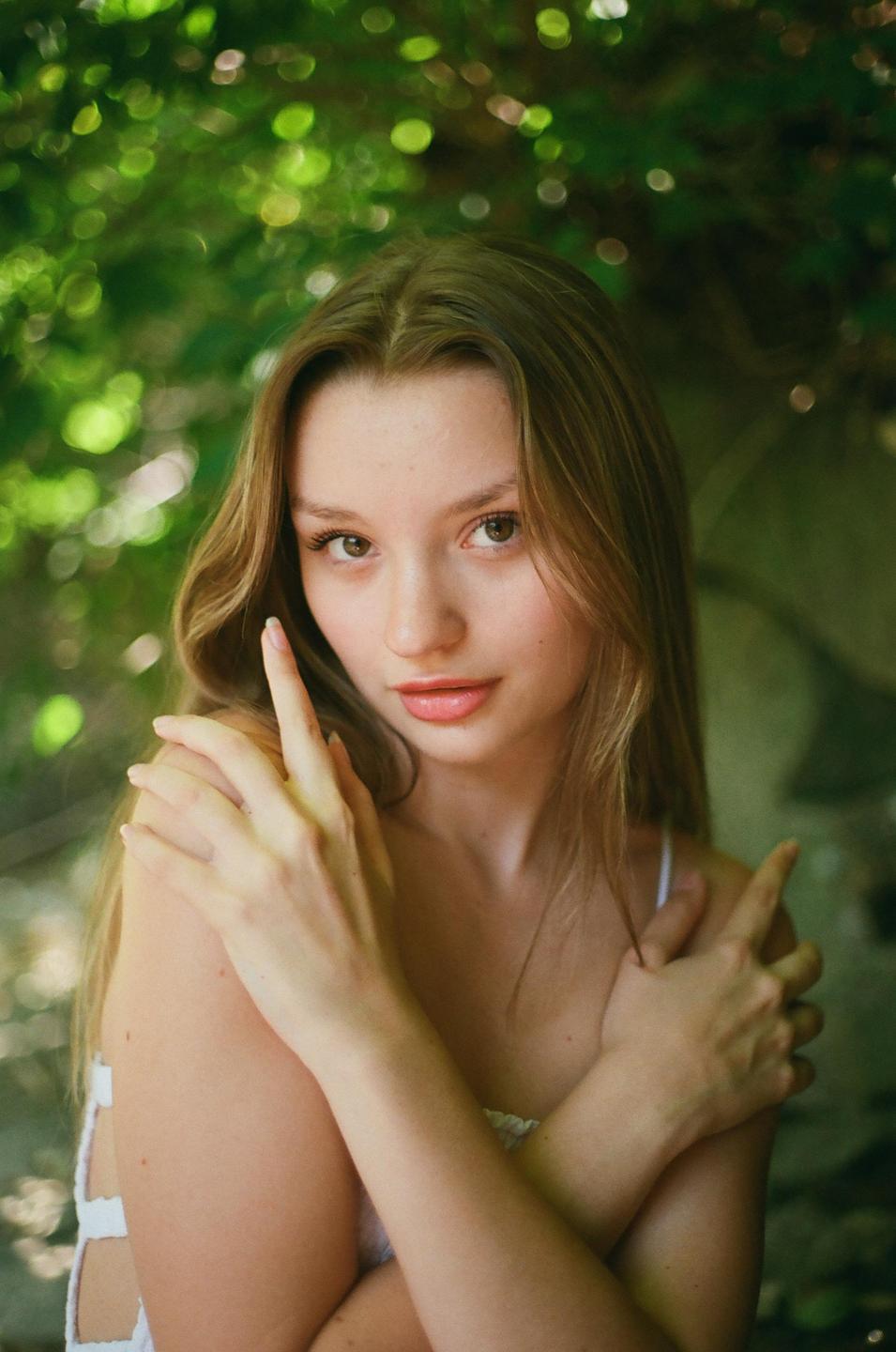 A woman with long hair poses with her arms crossed in front of a leafy green background.