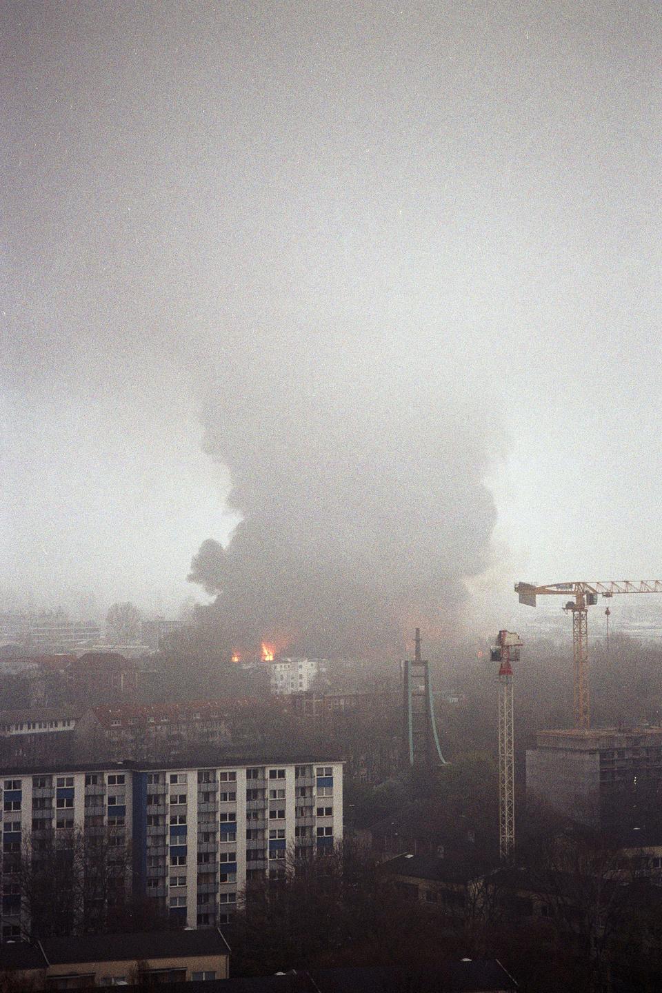 A large plume of smoke rises above a city with visible flames and construction cranes.