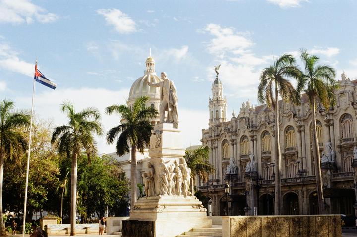 A statue of a man stands in front of a domed building and a large ornate building with palm trees and a Cuban flag.