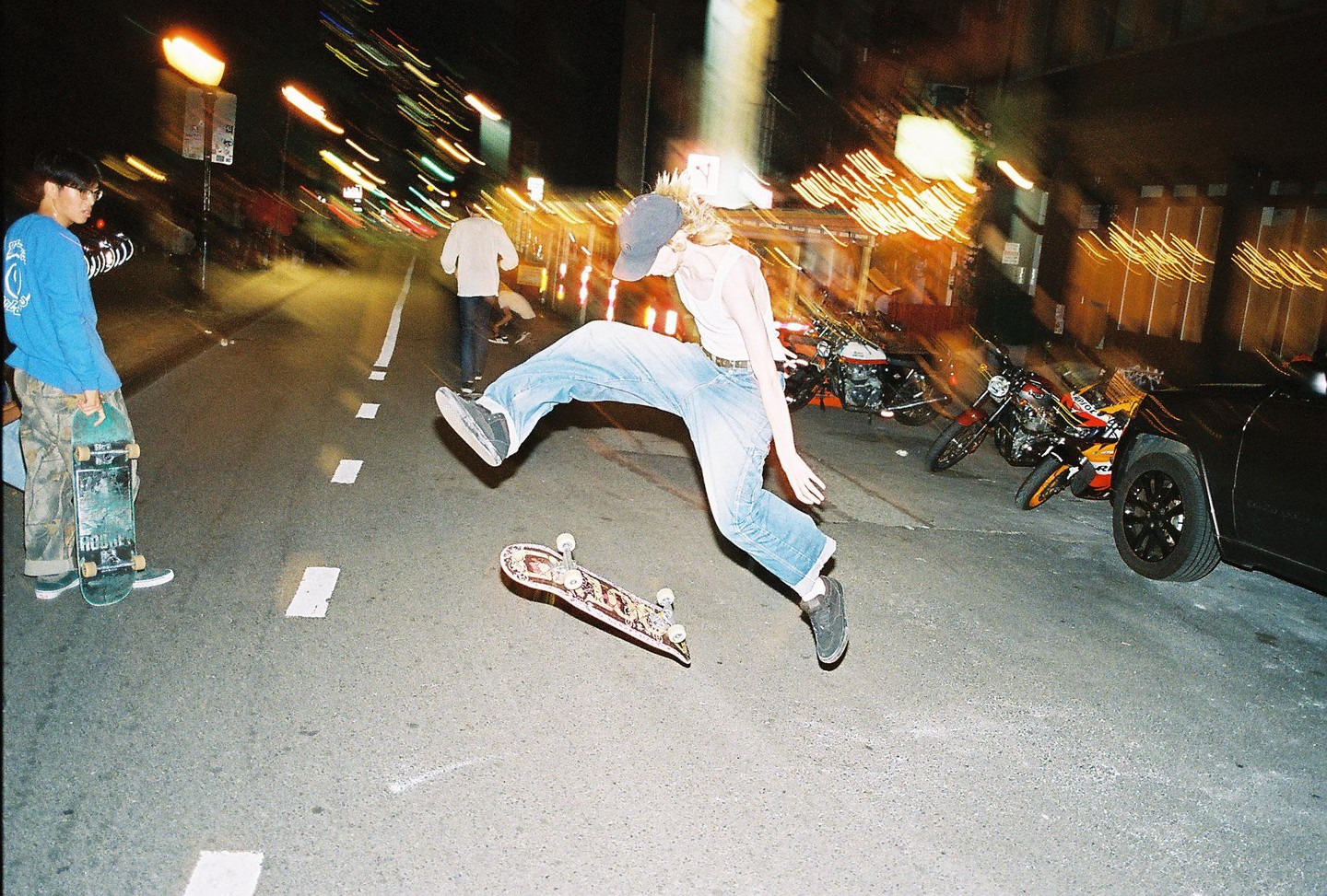 A young man in a tank top and jeans jumps over a skateboard on a street at night, with another young man holding a skateboard watching him.