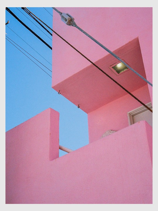 A bright pink building with power lines and a light fixture is set against a clear blue sky.