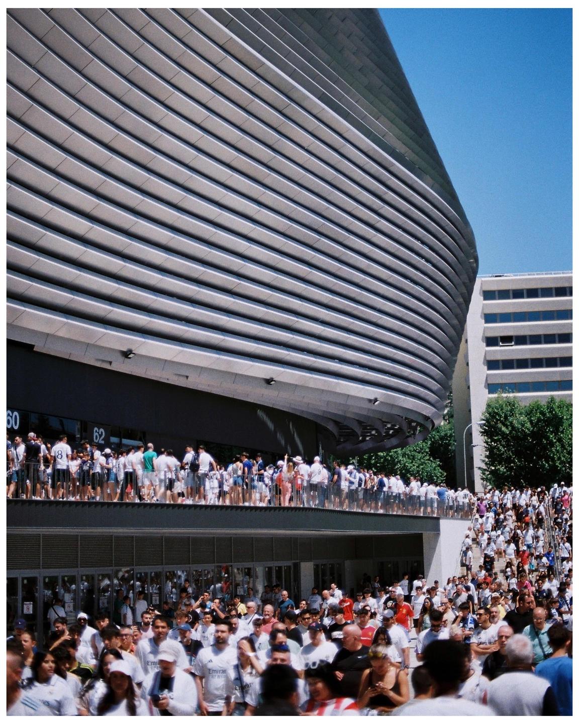 A large crowd of people gathers outside a modern stadium building under a clear blue sky.
