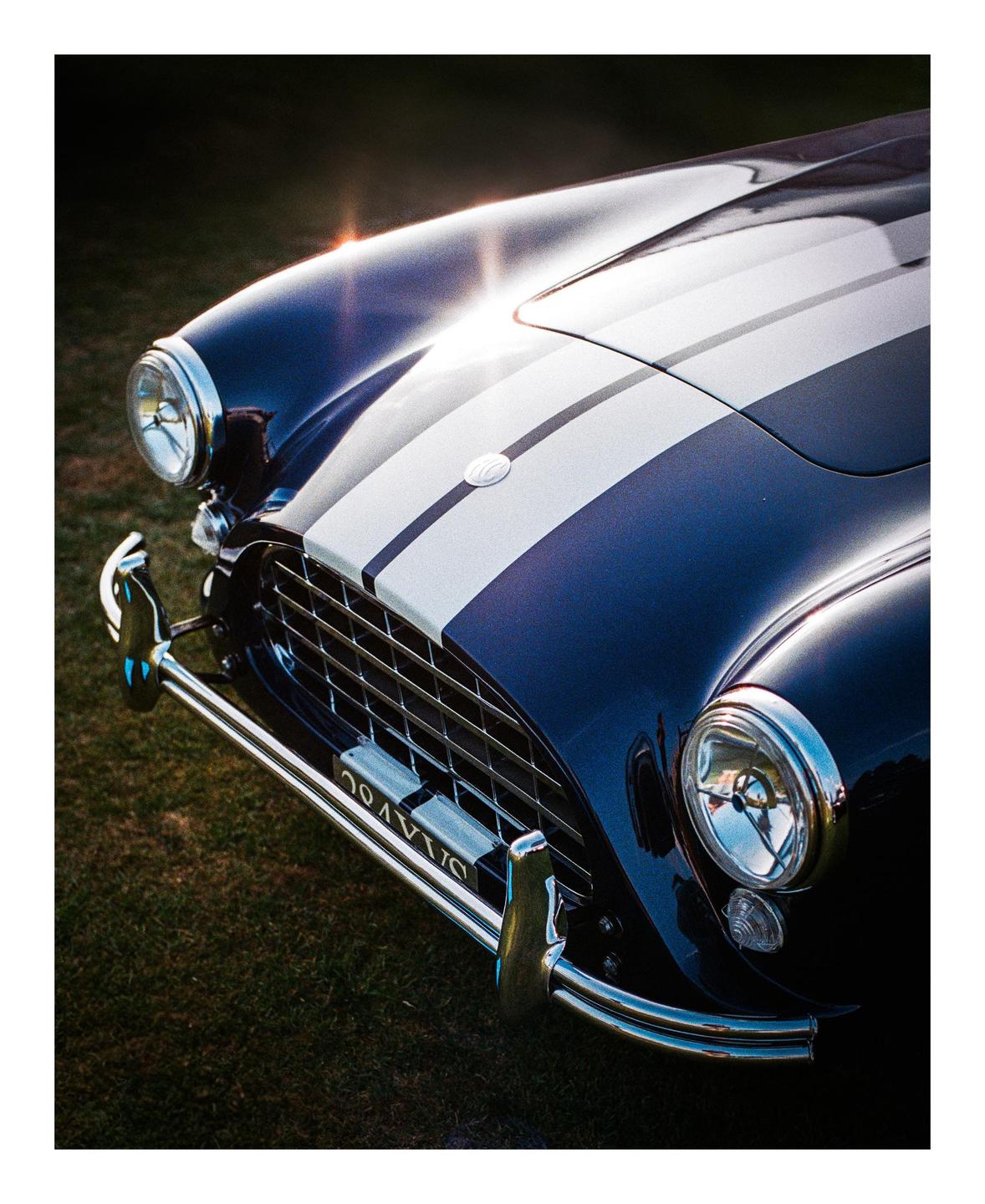 A blue vintage car with white racing stripes is parked on a grassy field.