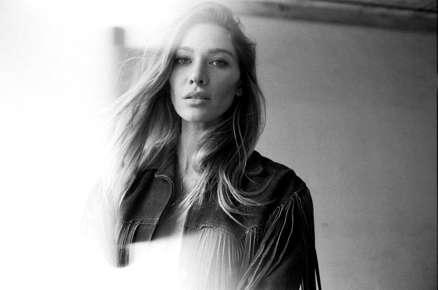A woman with long hair and a fringed jacket looks directly at the viewer.