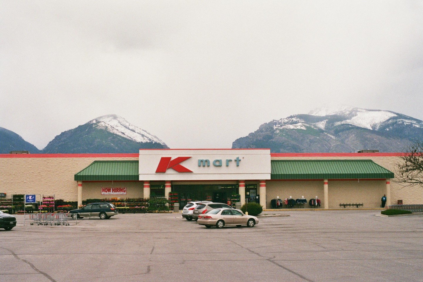 A Kmart store with a parking lot and cars is visible in front of snow-capped mountains under a cloudy sky.