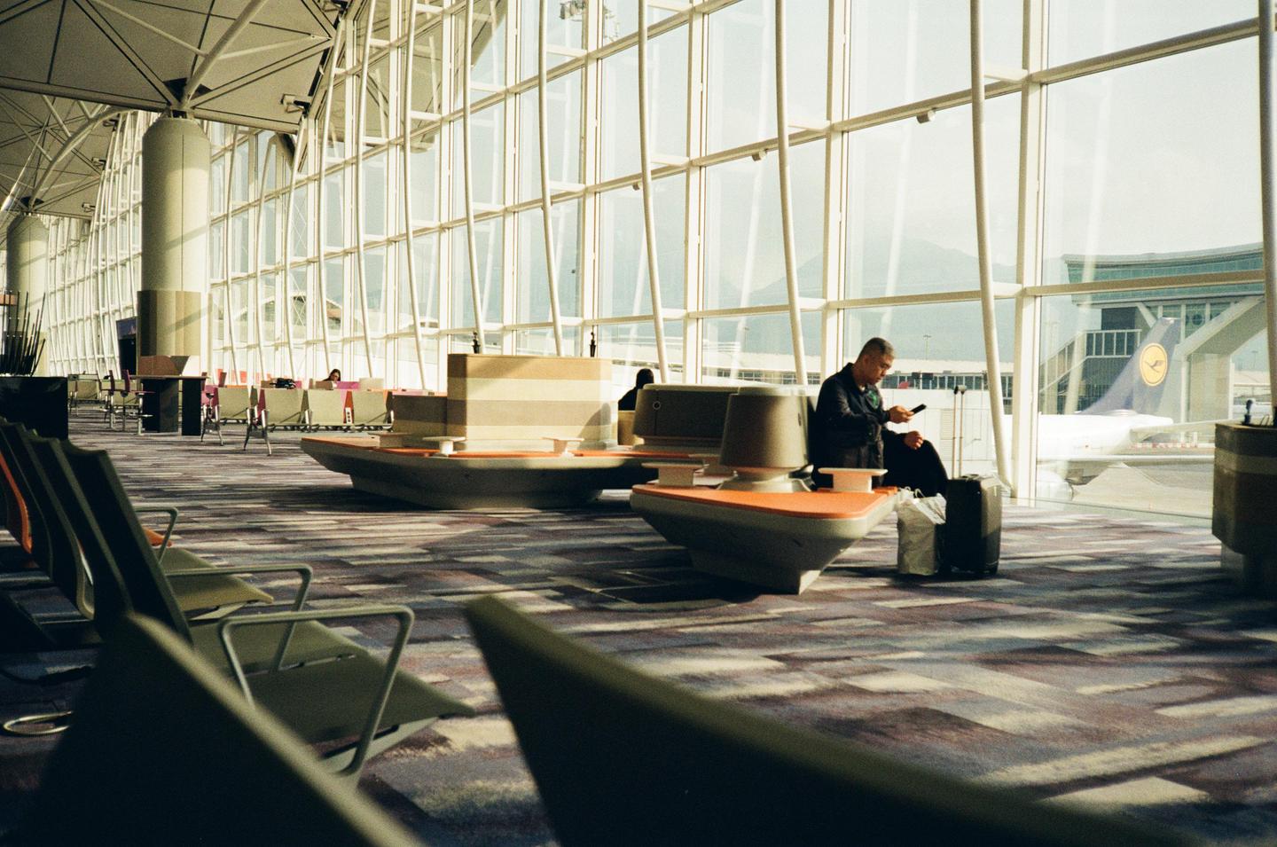 A man sits in an airport terminal, looking at his phone, with an airplane visible outside a large window.
