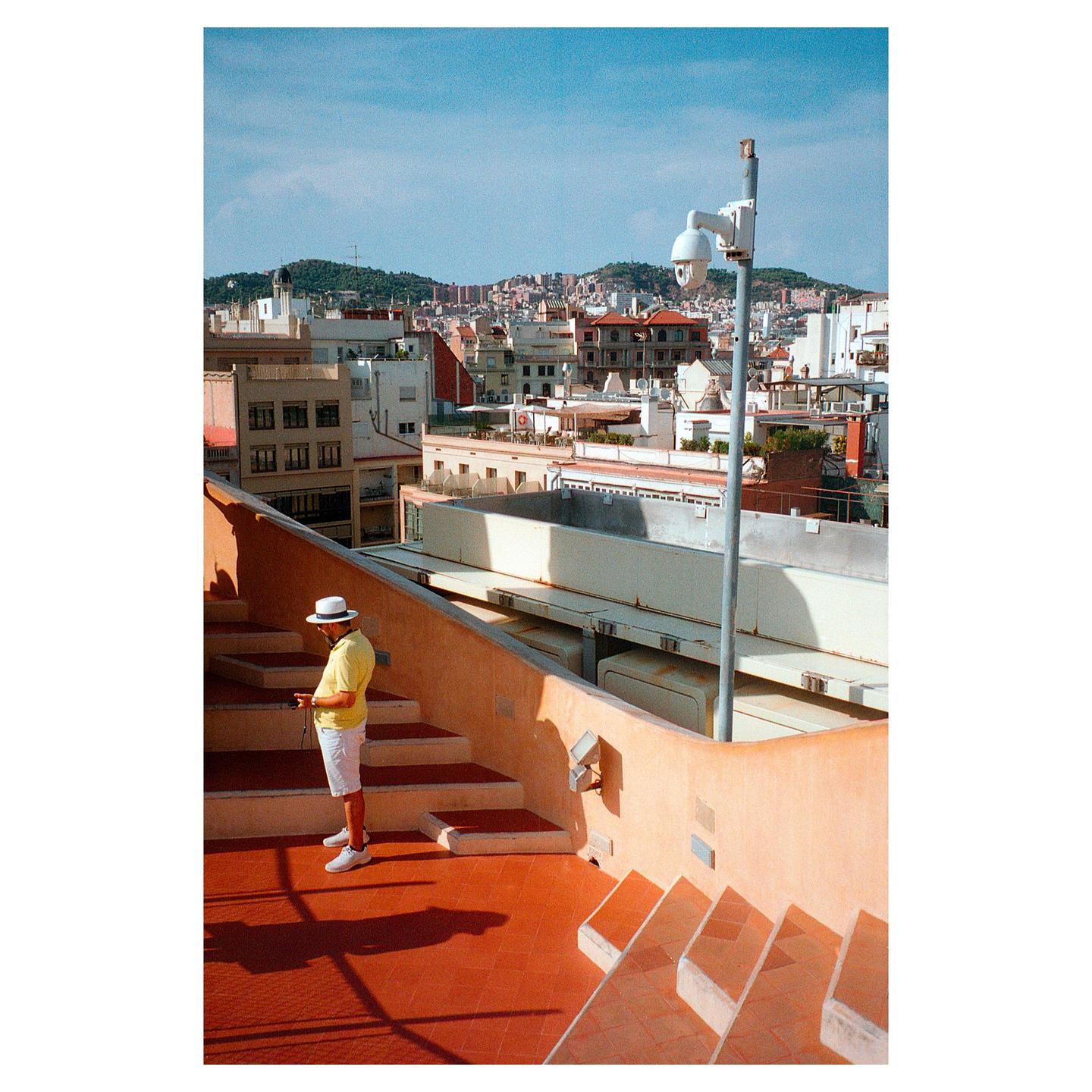 A man in a hat stands on a rooftop terrace overlooking a city.
