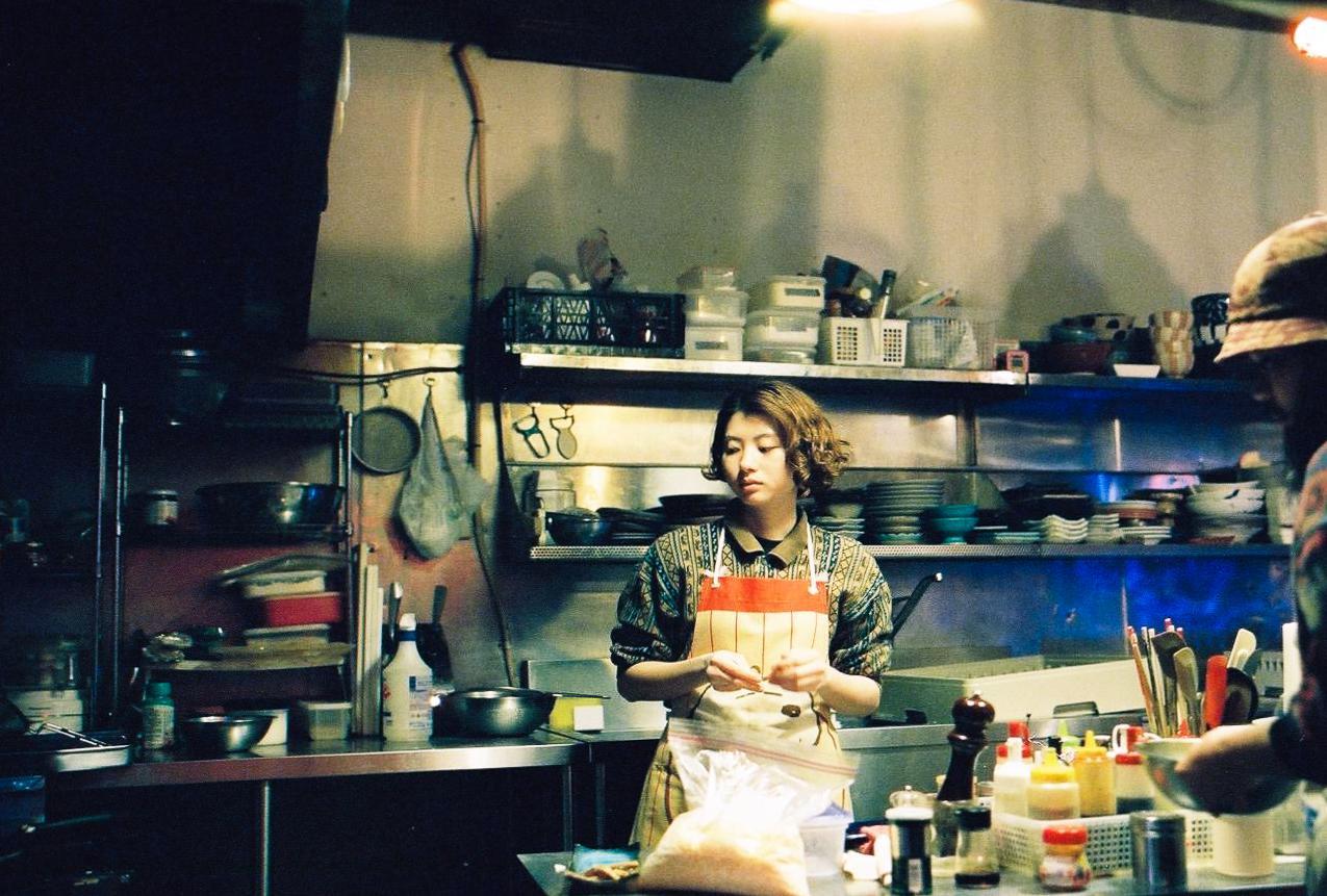 A woman wearing an apron stands in a busy kitchen, preparing food.
