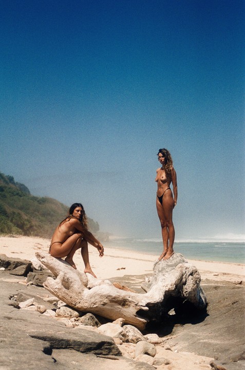Two women pose on rocks and driftwood on a sandy beach with a clear blue sky above.