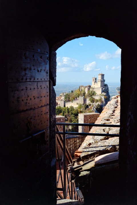 A view of a castle on a hill is framed by a dark archway and a wooden door.