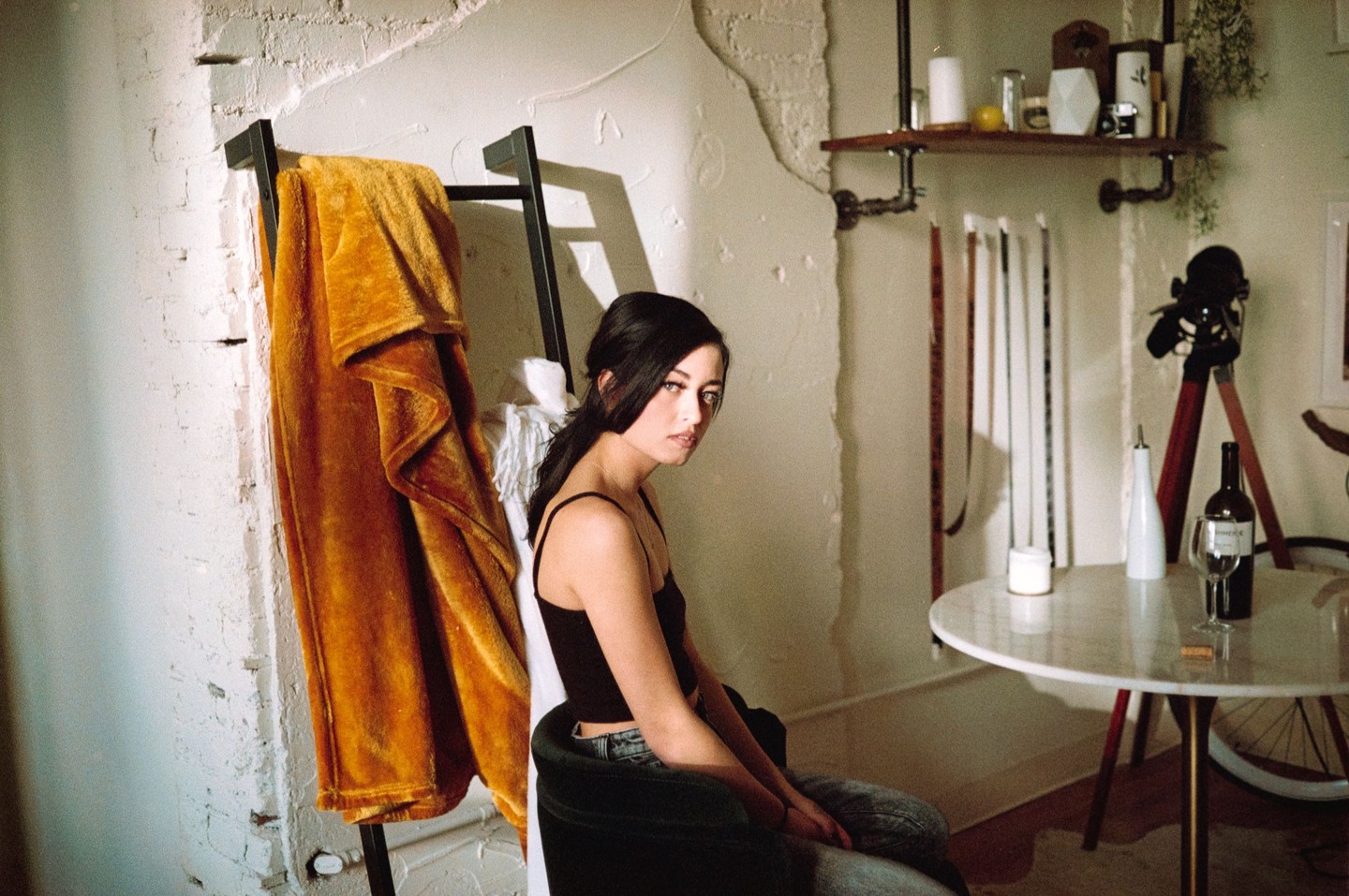 A woman sits in a chair next to a blanket draped over a ladder, with a table holding a bottle of wine and a glass to her right.