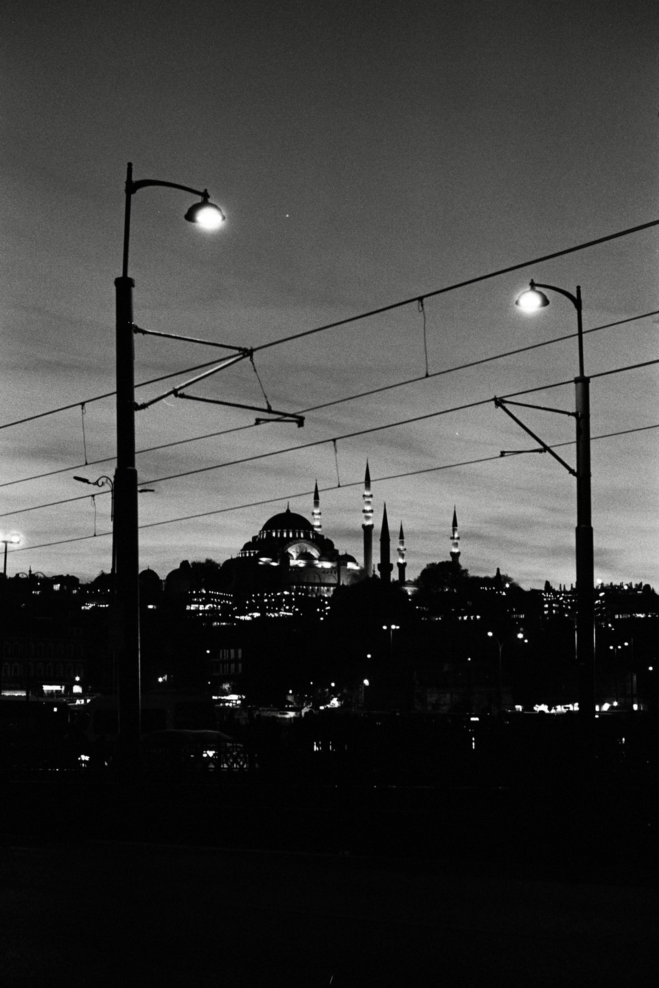 A black and white cityscape at night shows a mosque with multiple minarets and domes, illuminated against the dark sky, with streetlights and power lines in the foreground.