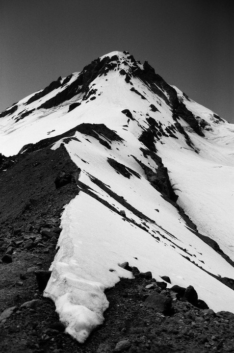 A snow-covered mountain peak rises sharply against a clear sky, with a rocky ridge leading up to it.