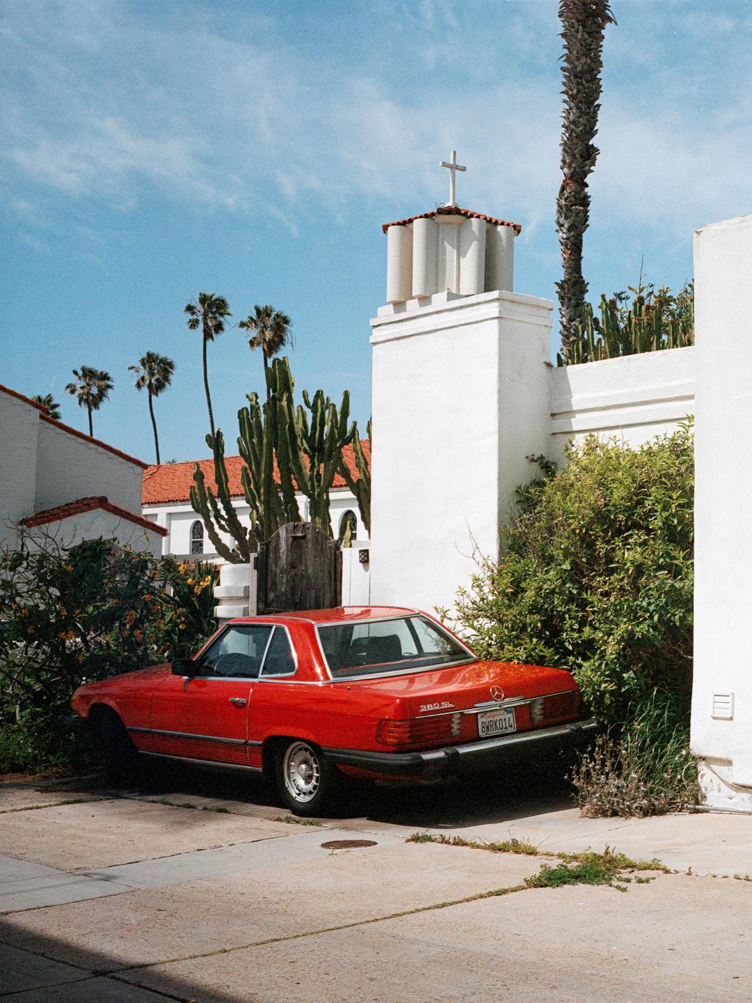 A red car is parked on a concrete driveway next to a white building with a cross on top, surrounded by palm trees and cacti.