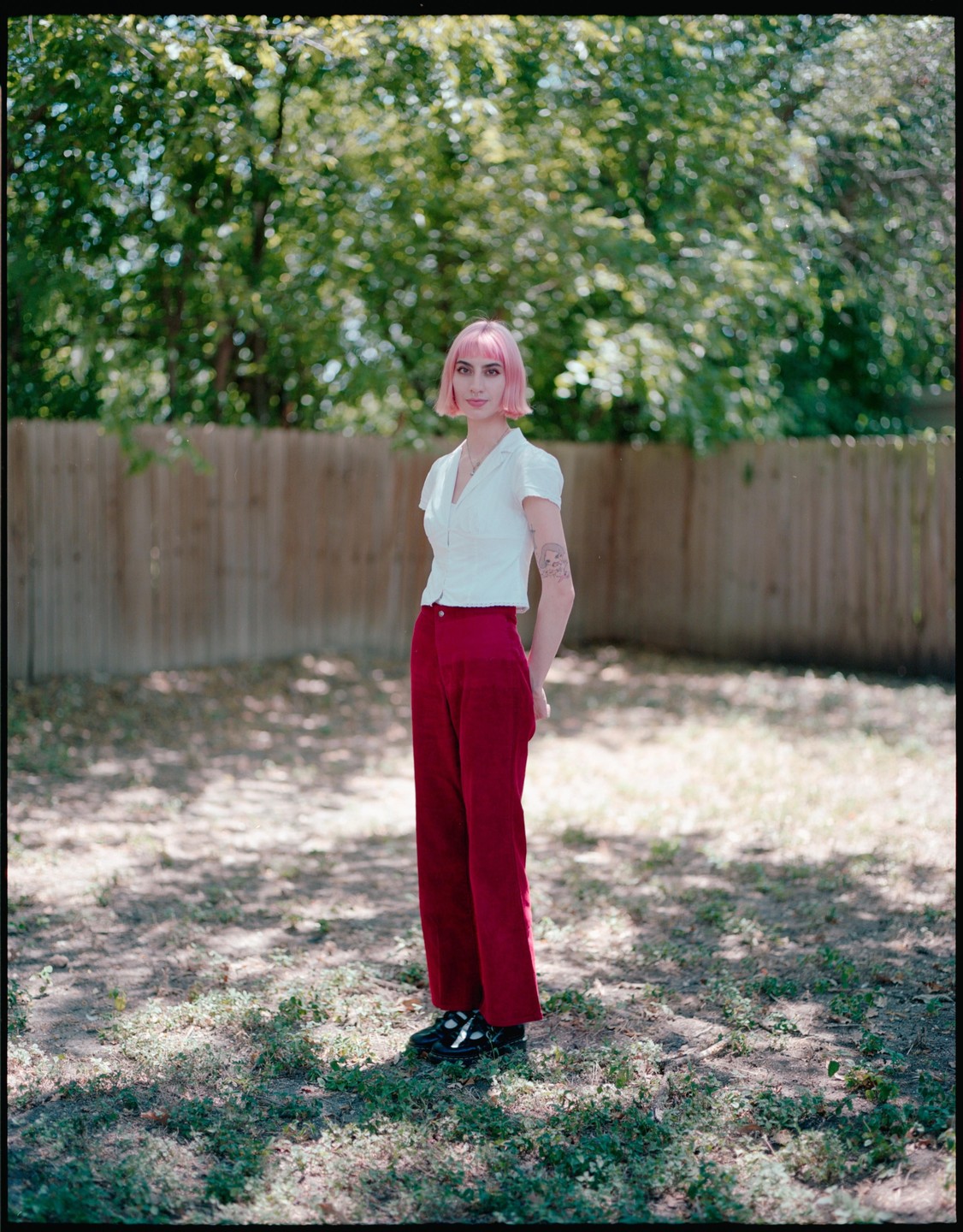 A woman with pink hair stands in a grassy yard with a wooden fence and trees in the background.