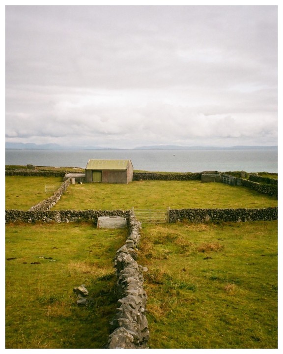 A small shed with a green roof stands in a green field enclosed by stone walls, with the ocean and a cloudy sky in the background.