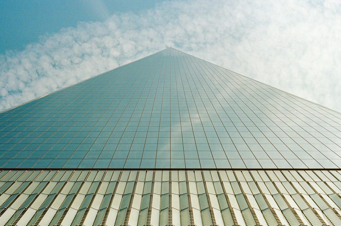 The glass facade of a skyscraper reflects the sky and clouds, with the building tapering towards a point.