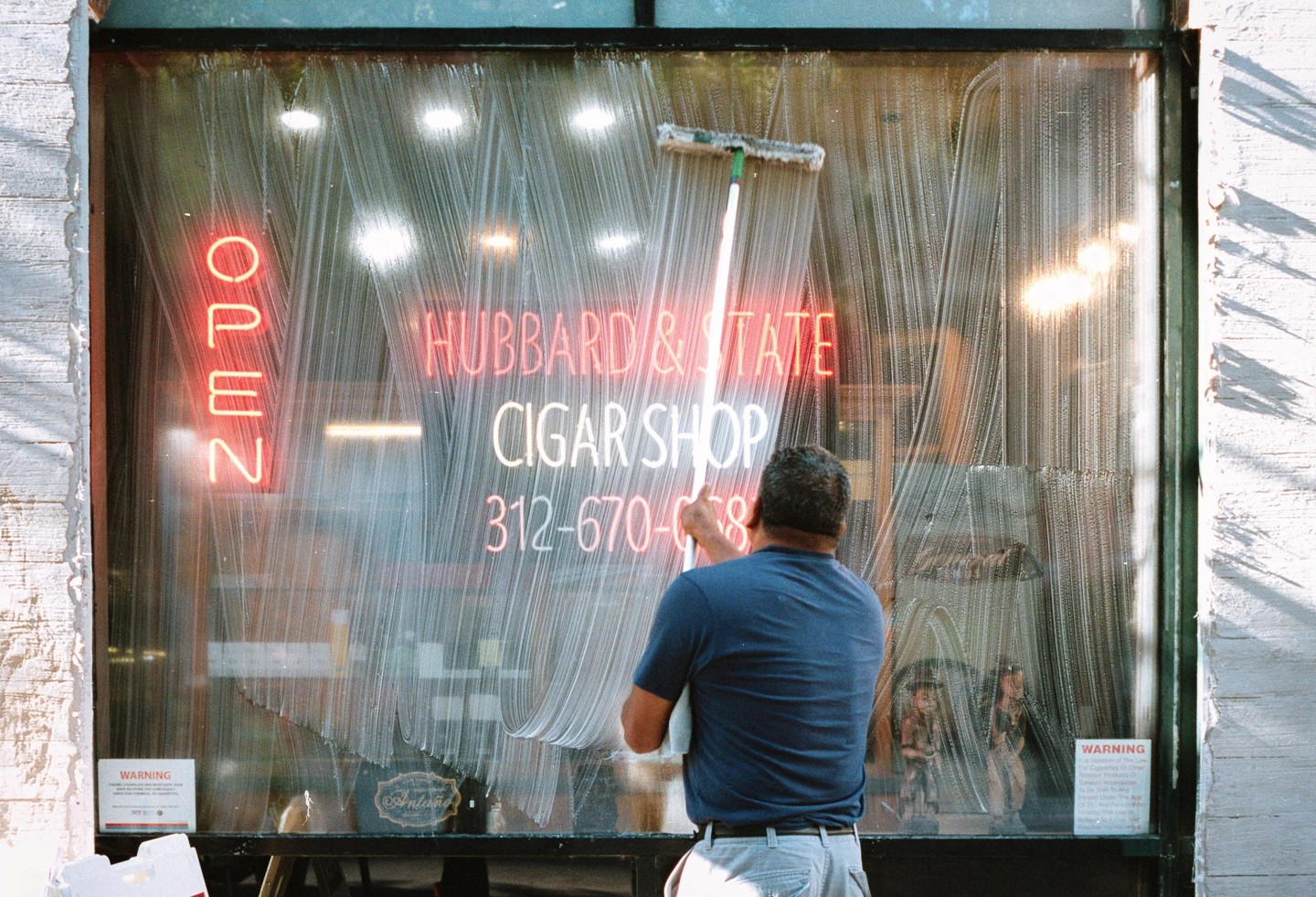 A man washes a storefront window with neon signs for a cigar shop.