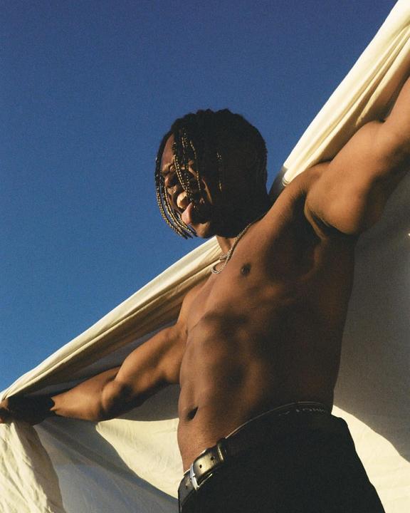 A man with braided hair and a necklace holds a white sheet against a clear blue sky.