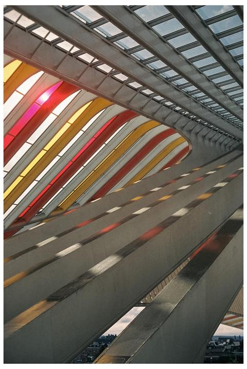 A modern train station features a glass roof with colorful red and yellow panels and strong diagonal lines.