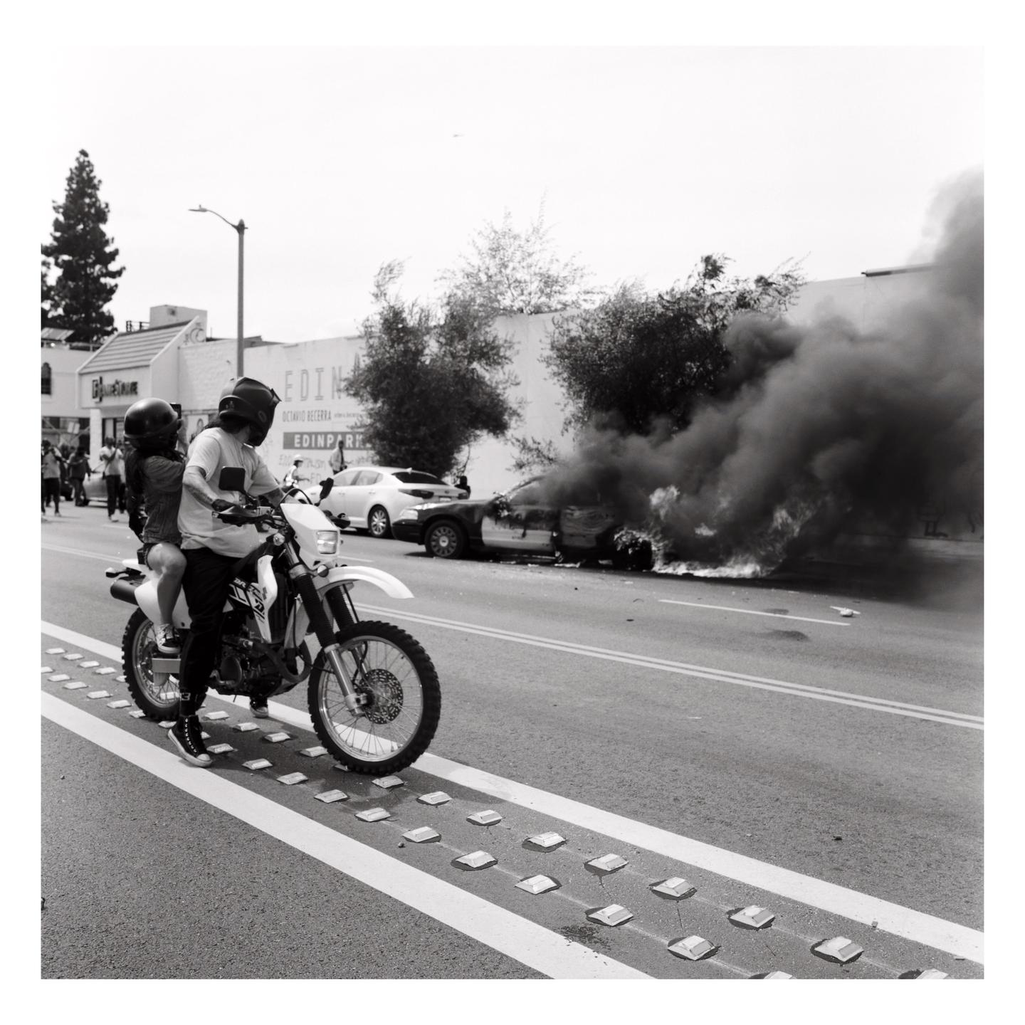 A man and woman on a motorcycle drive past a burning car on a street.