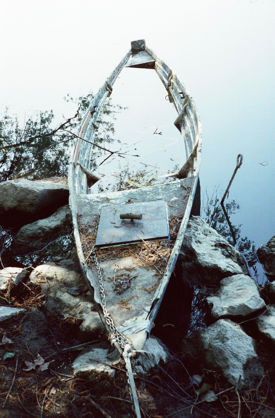 A small, old boat is nestled among rocks at the edge of a body of water, with reflections of trees visible in the water.