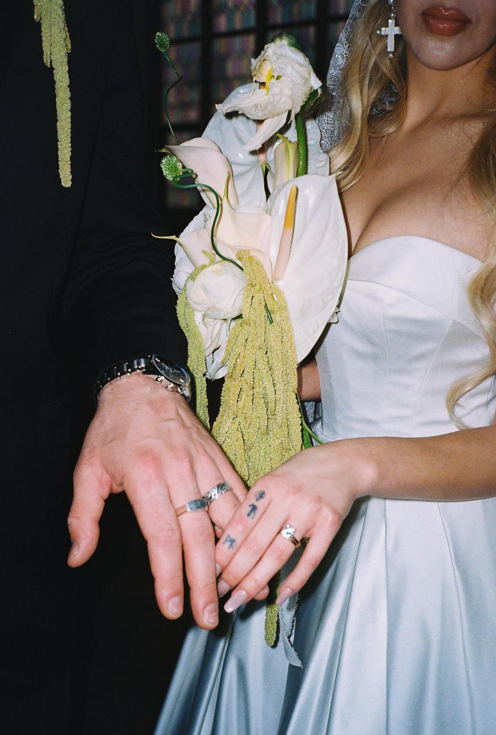 A couple holds hands, showing their rings and a wedding bouquet.
