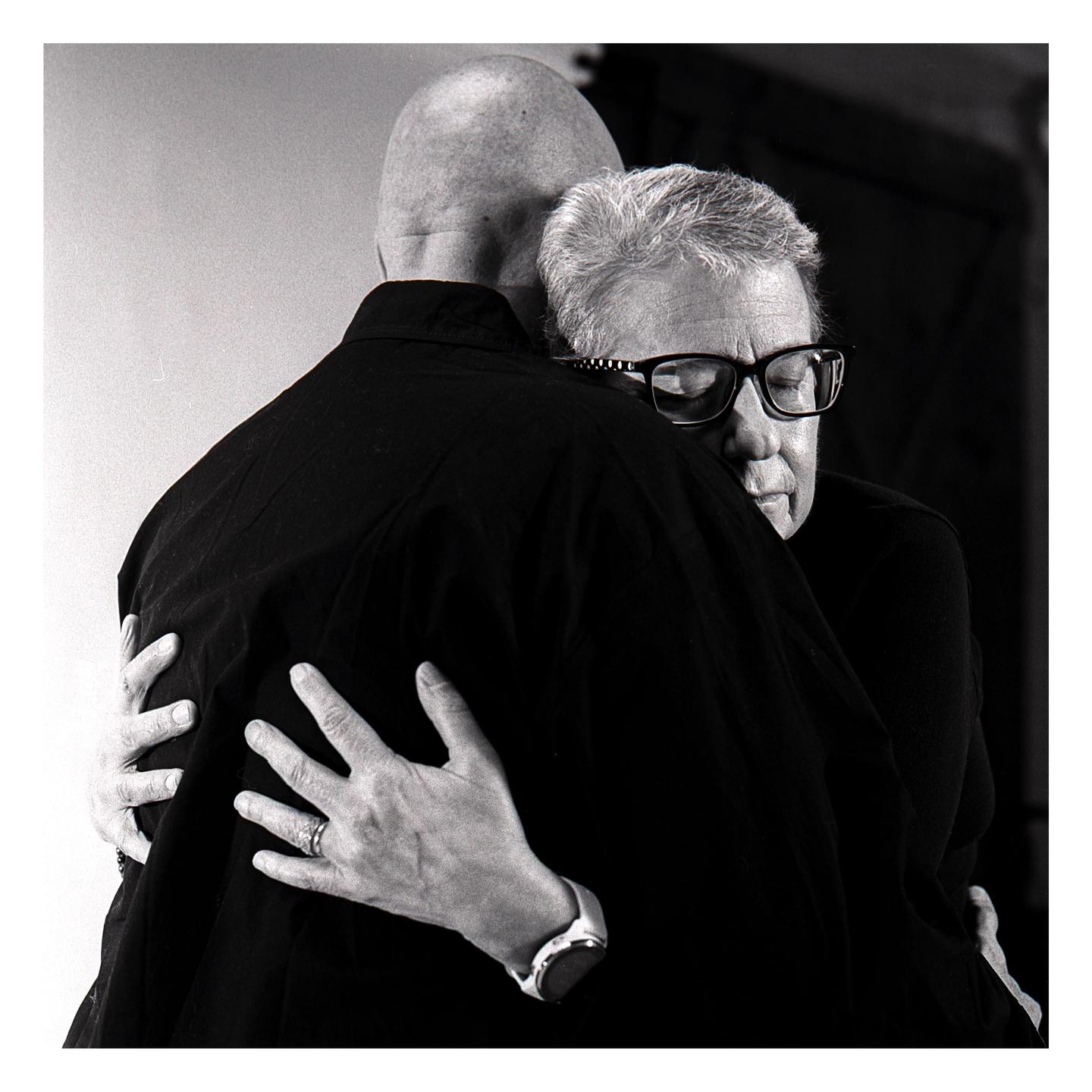 A man with glasses is embracing another man from behind.