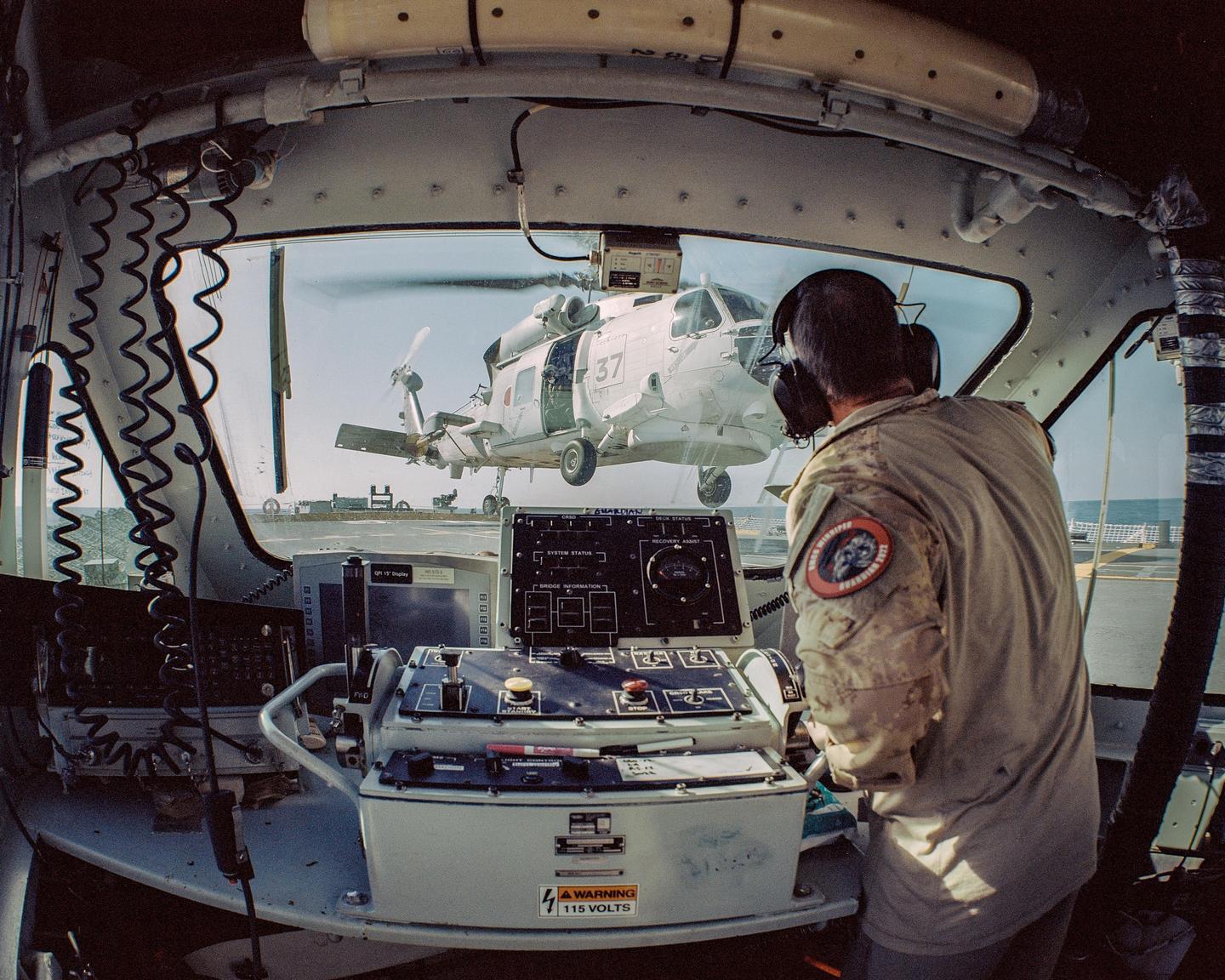 A man in a cockpit looks out at a helicopter hovering over a ship's deck.