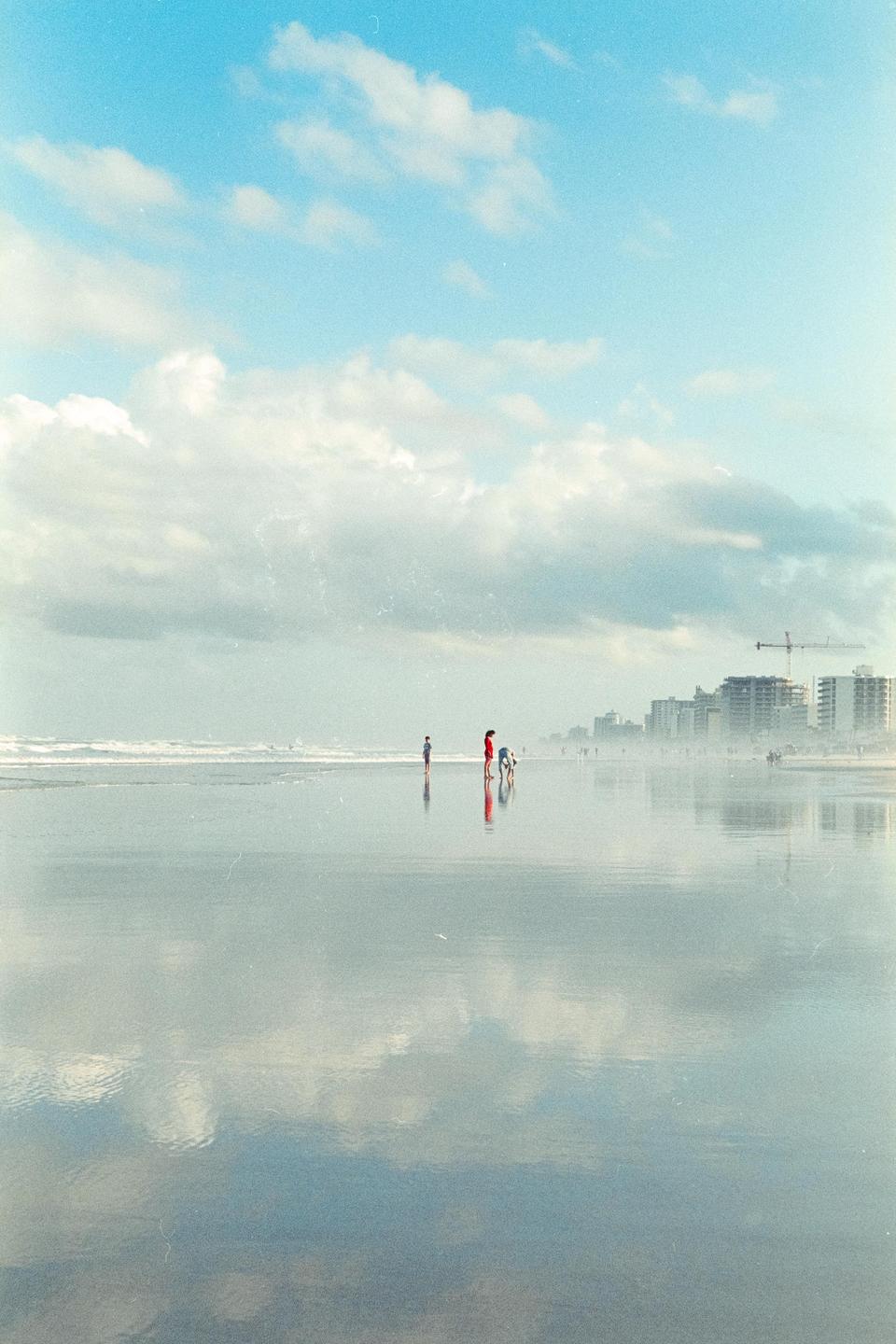 A group of people stand on a wet beach with buildings in the distance under a cloudy sky.