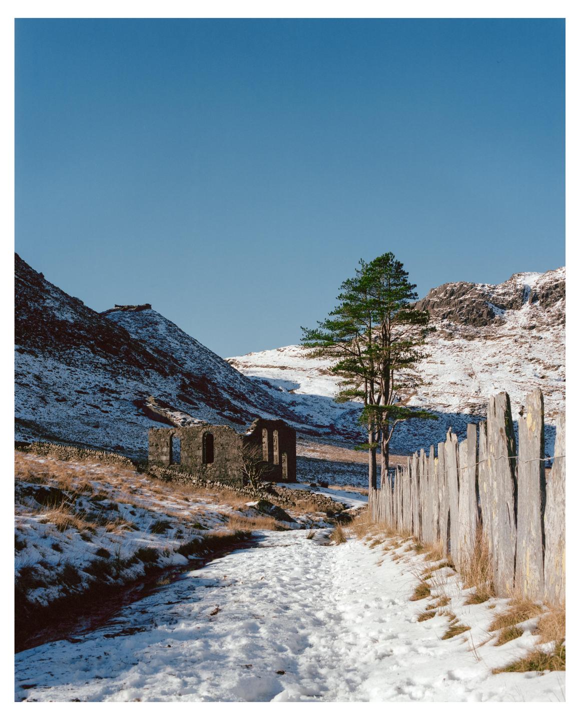 A snowy path leads to a ruined building in a mountain landscape with a wooden fence on the right.