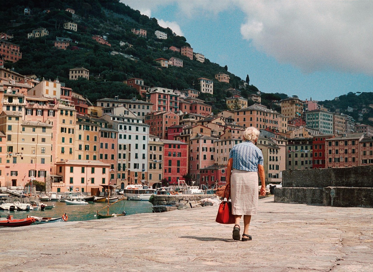 An elderly woman walks along a stone pier towards a harbor filled with boats and colorful buildings built into a hillside.