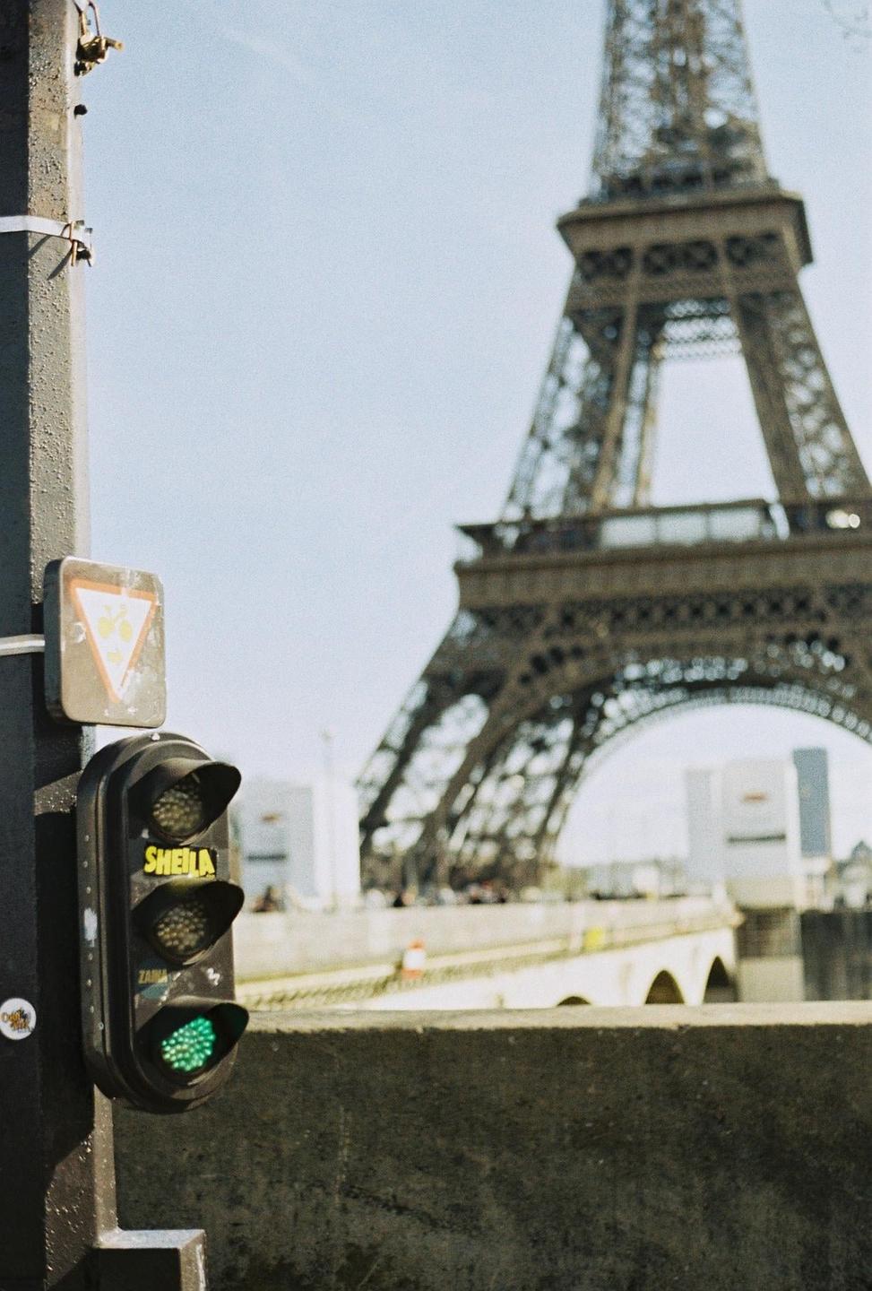 A traffic light with a green signal is in the foreground, with the Eiffel Tower and a bridge in the background.